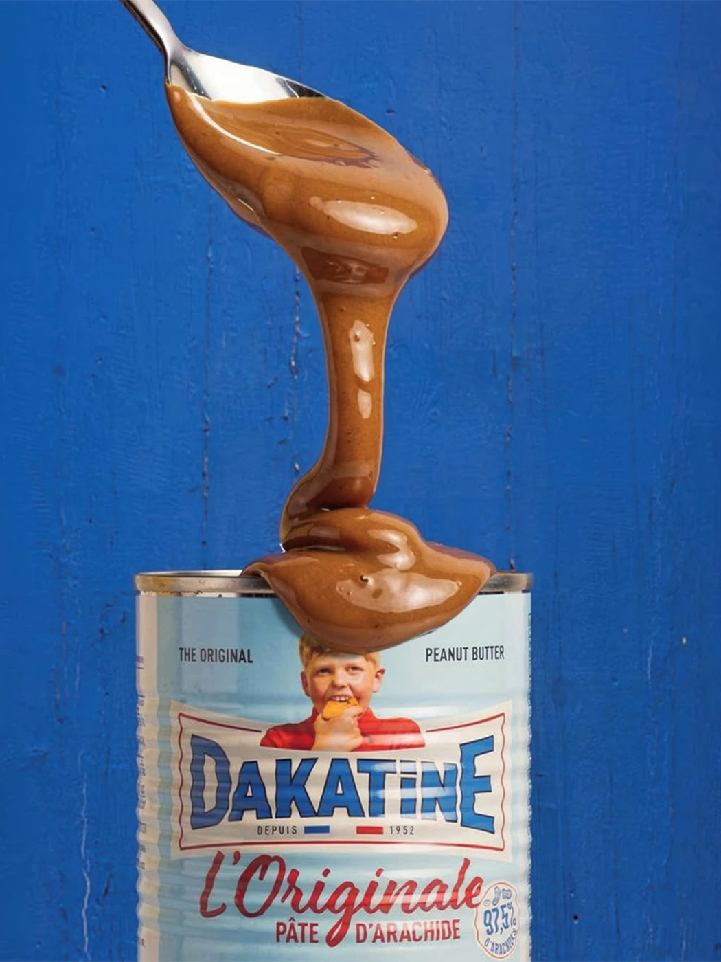 Cuillère versant la pâte d'arachide Dakatine sur une boîte L'Originale, fond bleu